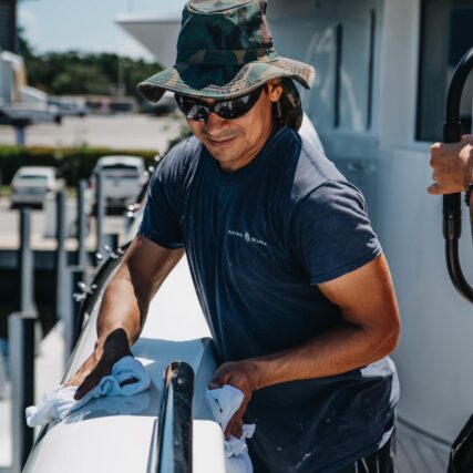 Marine worker wearing hat and sunglasses polishing a yacht’s deck on a sunny day.