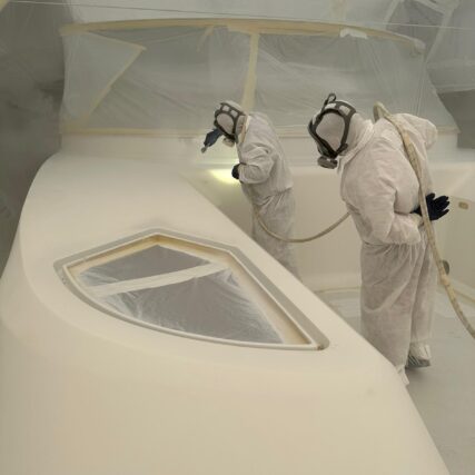 Two workers in protective suits painting or sanding the interior of a yacht hull.
