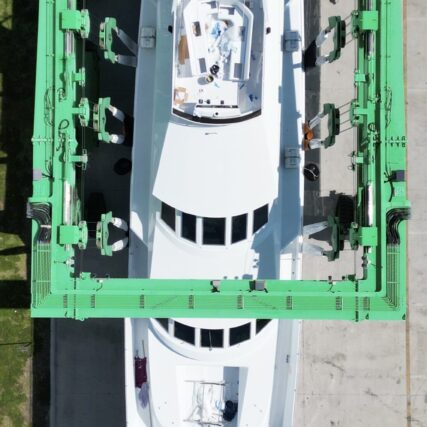Top-down view of a white yacht secured in a green boat lifting cradle at a shipyard.